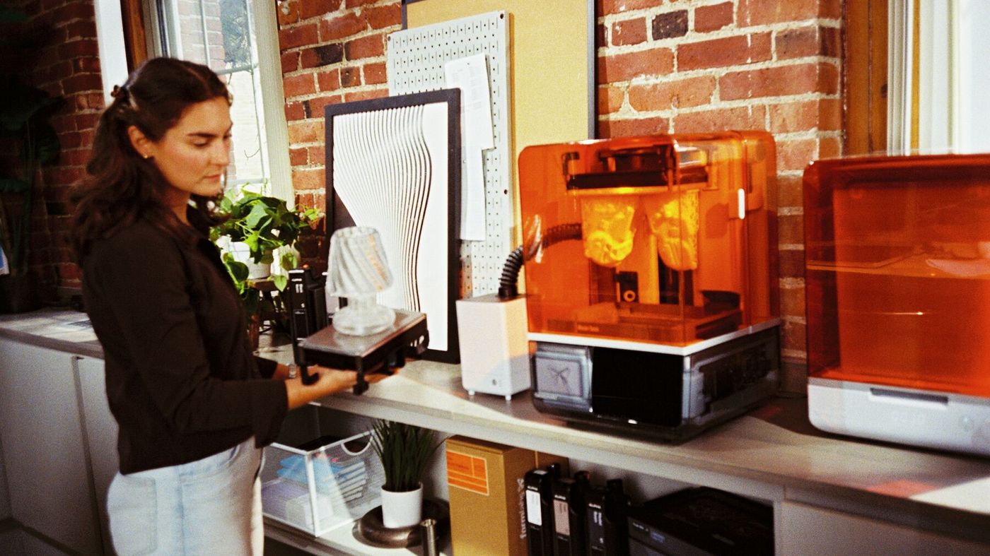 A person holding a finished print next to Form Studio 1, Form Studio 1 Wash+Cure and VOC Extractor in a sunny office