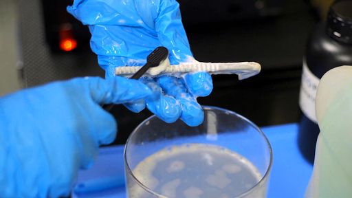 Blue-gloved hands scrub a resin part with a toothbrush over a cup