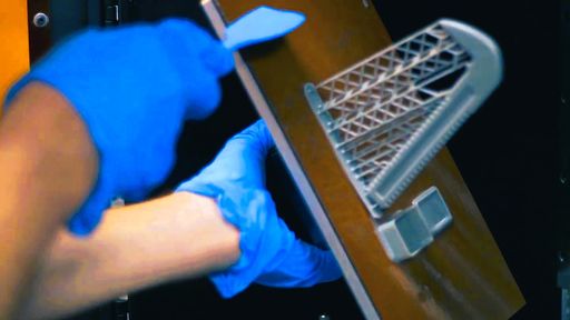 Blue-gloved hands pry a resin part off a tilted build plate with a spatula