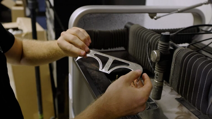 Hands holding a cut aluminum bracket beside a waterjet cutter